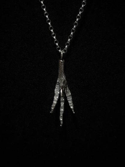 A solid silver casting of a pigeon claw hangs against a black background. This is a pendant and hung on a silver chain.