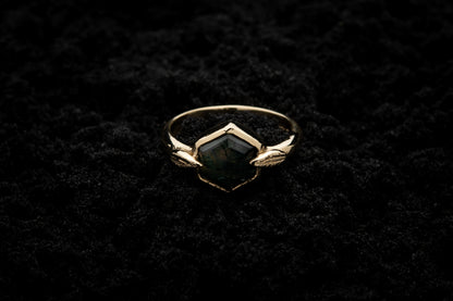 A leafy engagement ring rests on black sand and is lit in soft, glowy light.
A luscious hexagonal moss agate is set in a solid gold setting with a slim leaf on either side, folded slightly over the stone.