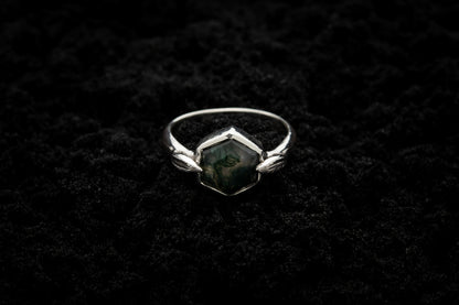 A leafy engagement ring rests on black sand and is lit in soft, glowy light.
A luscious hexagonal moss agate is set in a white gold setting with a slim leaf on either side, folded slightly over the stone.