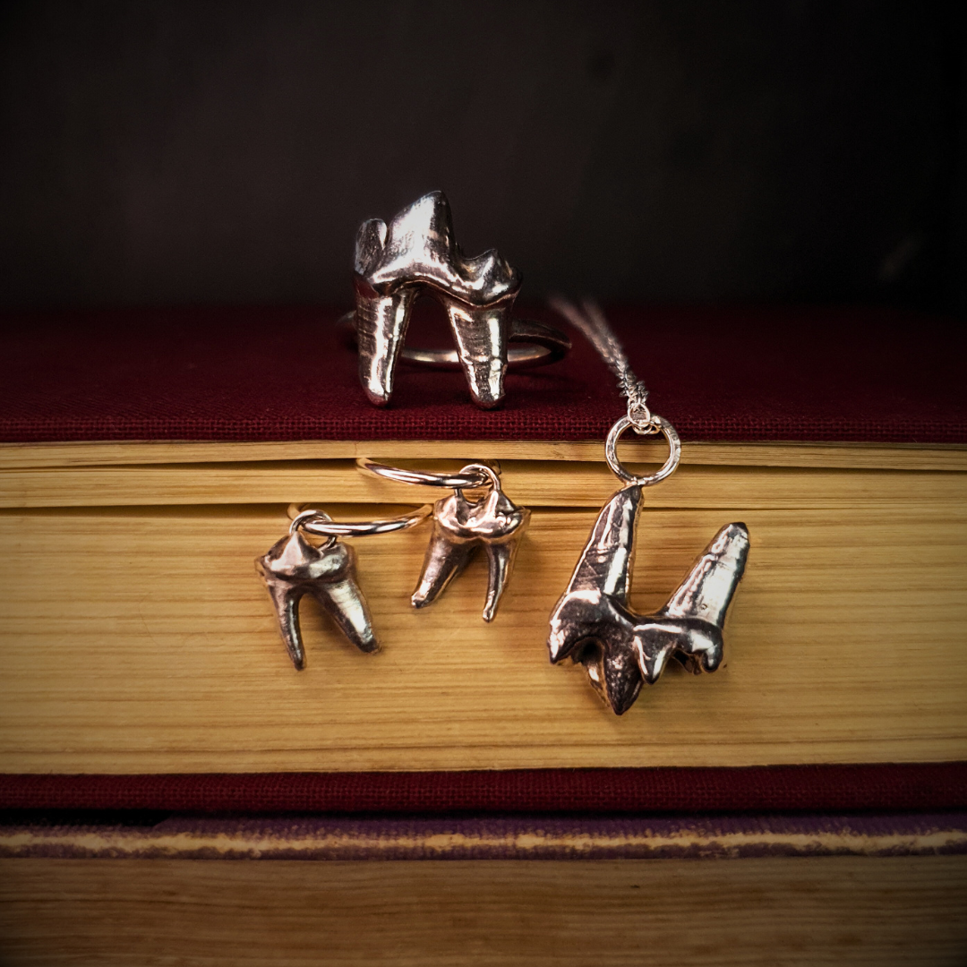 A sterling silver fox tooth jewellery sits atop a cloth-bound red book while a fox tooth necklace hangs over the edge. A pair of fox tooth hoop earrings is held between the aged pages.