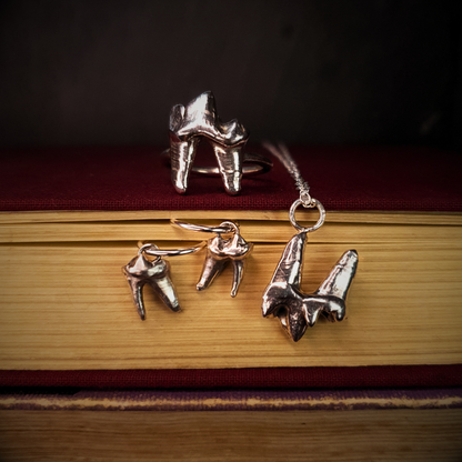 A sterling silver fox tooth jewellery sits atop a cloth-bound red book while a fox tooth necklace hangs over the edge. A pair of fox tooth hoop earrings is held between the aged pages.