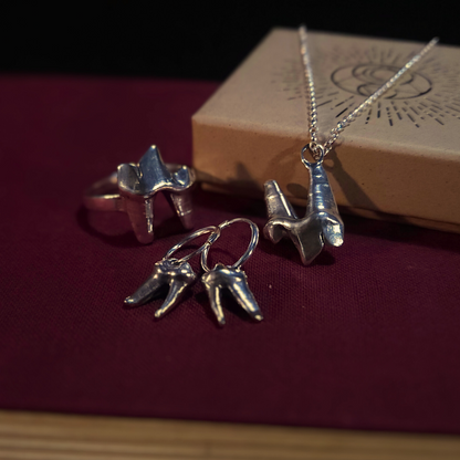 A set of sterling silver fox tooth jewellery sit atop a cloth-bound red book and next to a natural cardboard jewellery box. The set is comprised of a ring, earrings and a necklace.
