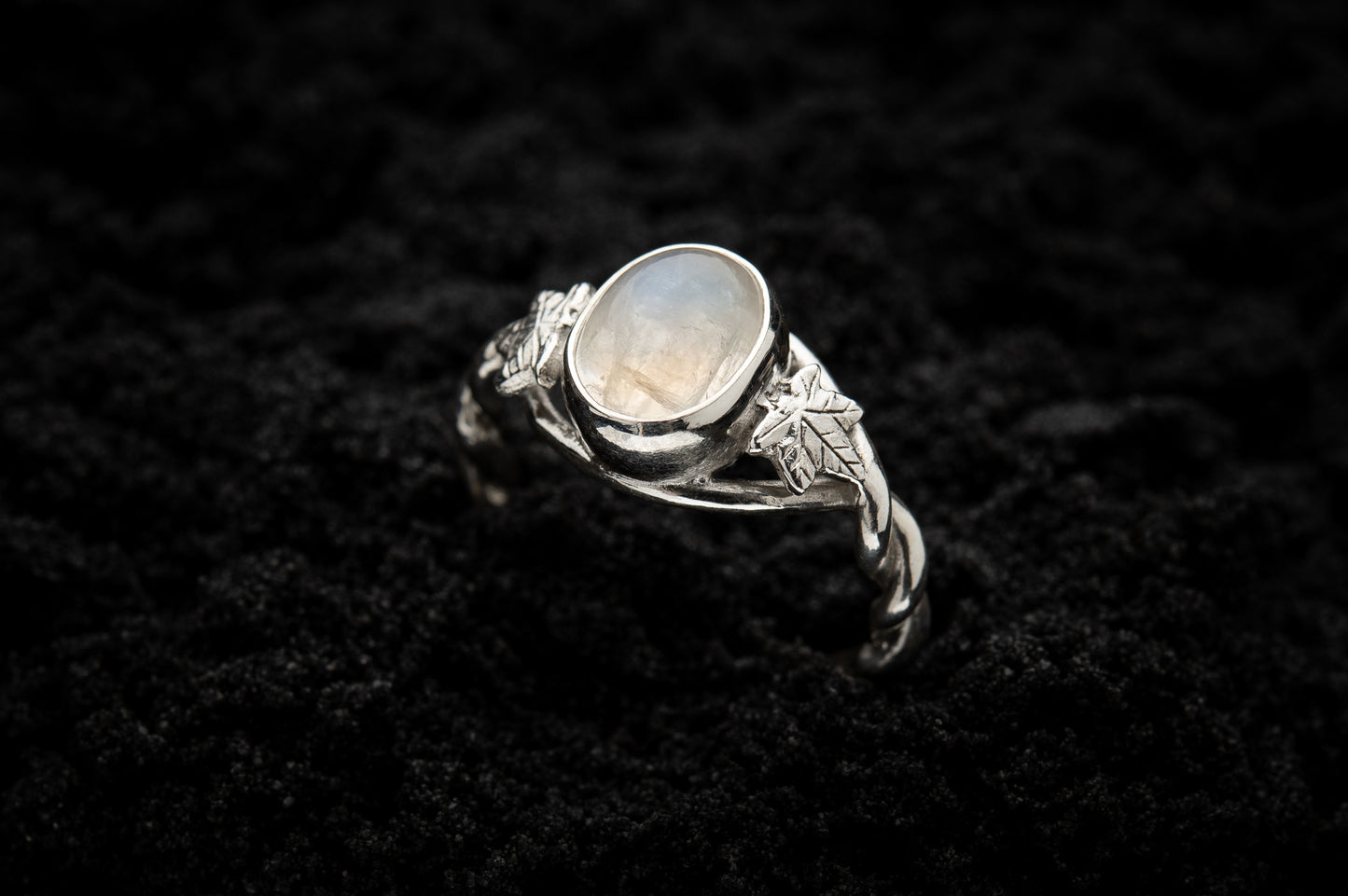A botanical engagement ring made from rainbow moonstone and white gold sits, half submerged, in black sand. It features a twisted band and an ivy leaf on each shoulder. The moonstone shines gently in the camera flash, teasing it's bluey rainbow hues.
