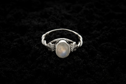 A botanical engagement ring made from rainbow moonstone and 9 carat white gold lays atop black sand. It features a twisted band and an ivy leaf on each shoulder. The moonstone shines gently in the camera flash, teasing it's bluey rainbow hues.