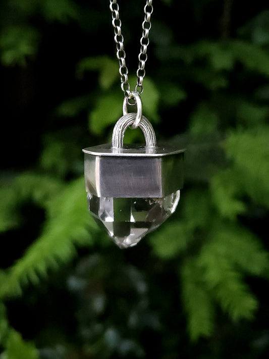 A chunky sterling silver pendant with a faceted garden quartz point, hanging on a belcher chain. Photographed outdoors against a blurred green foliage background.