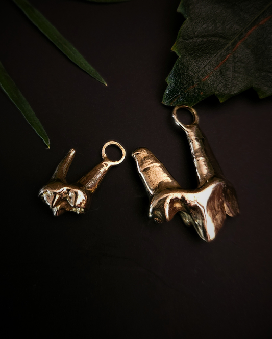 2 yellow gold pendants made from the casting of a baby fox tooth sits on a black background with a few pieces of green foliage around them. A small one is on the left and a large one is on the left. The photograph is taken from the top down. This is a high end taxidermy style pendant for fans of gothic jewellery, the macabre, unusual and taxidermy.