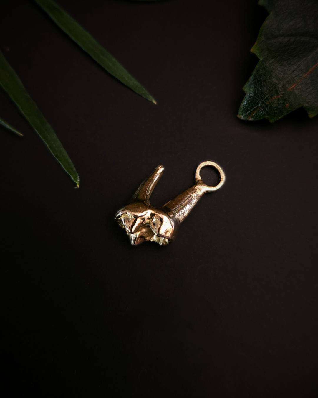 A small, yellow gold pendant made from the casting of a baby fox tooth sits on a black background with a few pieces of green foliage around it. This is a luxury gothic pendant for fans of alternative jewellery, the witchy, unusual and folkloric.