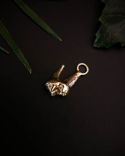A small, yellow gold pendant made from the casting of a baby fox tooth sits on a black background with a few pieces of green foliage around it. This is a luxury gothic pendant for fans of alternative jewellery, the witchy, unusual and folkloric.