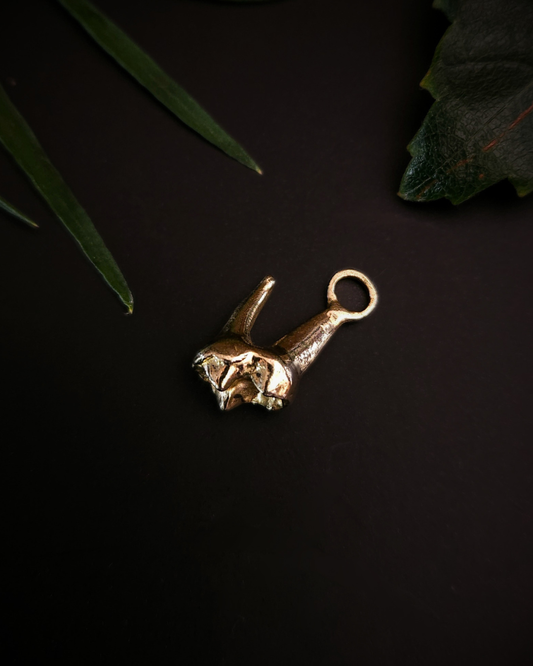 A small, yellow gold pendant made from the casting of a baby fox tooth sits on a black background with a few pieces of green foliage around it. This is a luxury gothic pendant for fans of alternative jewellery, the witchy, unusual and folkloric.