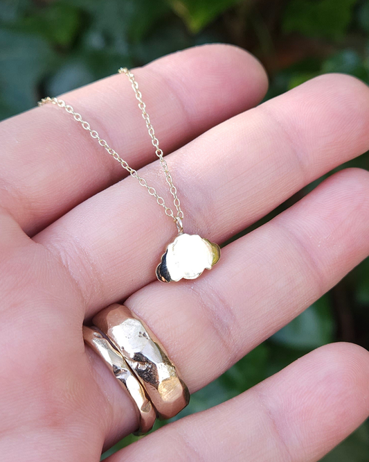 A shiny solid gold cloud necklace is held in an open hand to show its dainty size. The hand is also wearing 2 gold wedding rings. There is green foliage in the background.