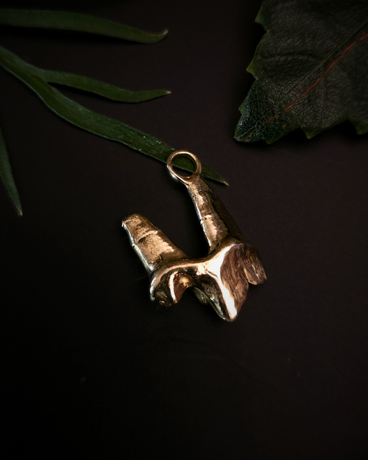A solid yellow gold pendant, made from the casting of a fox tooth, sits on a black background with a few pieces of green foliage around. The photograph is taken from the top down. This is a chunky and luxury taxidermy style pendant for fans of gothic jewellery, the macabre, unusual and taxidermy.