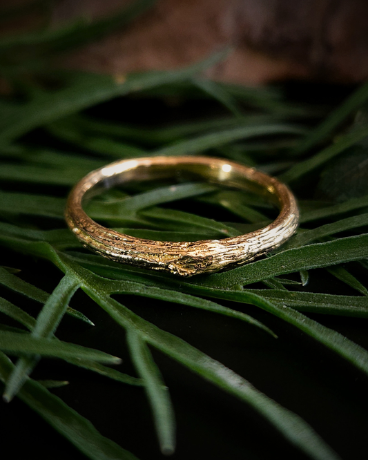A luxuriously textured 9ct gold ring with a natural bark-like surface, resting on green foliage against a dark background. There is a small wood-knot in the centre of the ring.