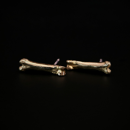 A pair of witchy 9 carat gold bone stud earrings laying on their side on a solid black background.
