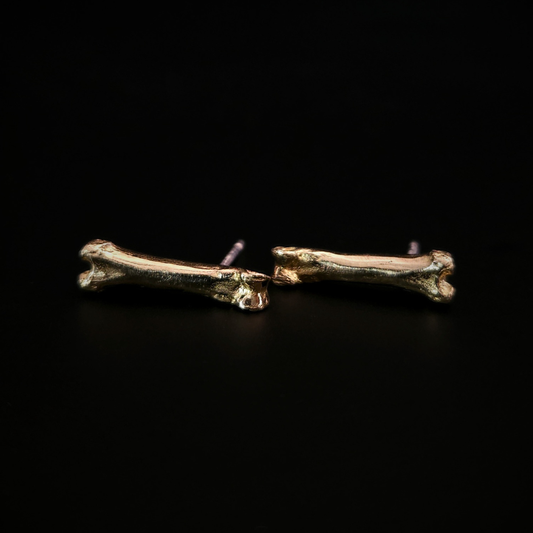 A pair of witchy 9 carat gold bone stud earrings laying on their side on a solid black background.