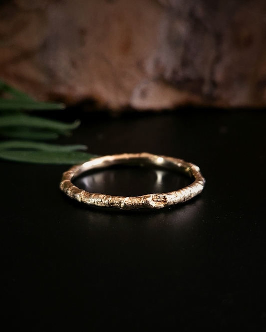 A 9 carat gold ring made from the casting of a thin twig sits on a black surface with some brown bark and green foliage out-of-focus in the background. The inside of the ring is nice and smooth for comfort and the light reflects from it in a soft golden glow.