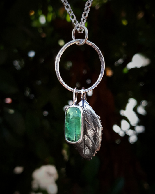 Silver necklace with a solid silver leaf and mint green stone pendant hang from a silver circlet against a blurred natural background