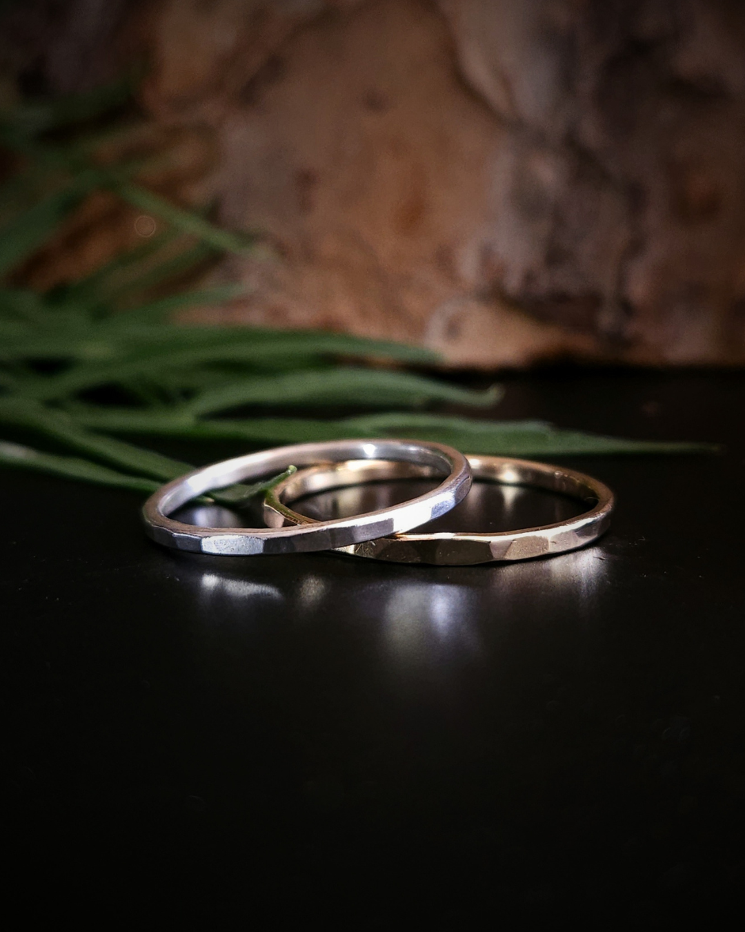 A hammered, dainty, sterling silver and 9ct yellow gold sit on black surface with a natural bark and foliage background. Their smooth, shiny, hammered edges reflect the light beautifully.