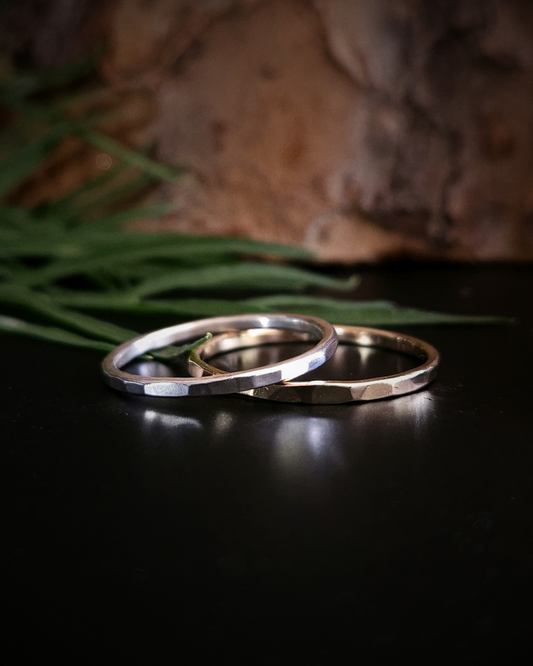 A hammered, dainty, sterling silver and 9ct yellow gold sit on black surface with a natural bark and foliage background. Their smooth, shiny, hammered edges reflect the light beautifully.