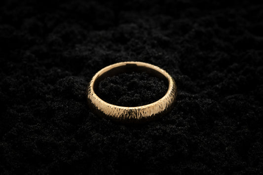 A thick yellow gold wedding ring sits in black sand to contrast the warm luscious glow. There are hammered striations around the band like the suns rays.