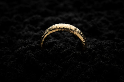 A thick yellow gold wedding ring sits in black sand to contrast the warm luscious glow. There are hammered striations around the band like the suns rays. It is half submerged in the sand and breaks through the surface like a golden rainbow.