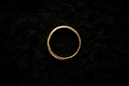 A thick yellow gold wedding ring sits in black sand to contrast the bright, warm, luscious glow. There are hammered striations around the band like the suns rays. It is photographed from above.