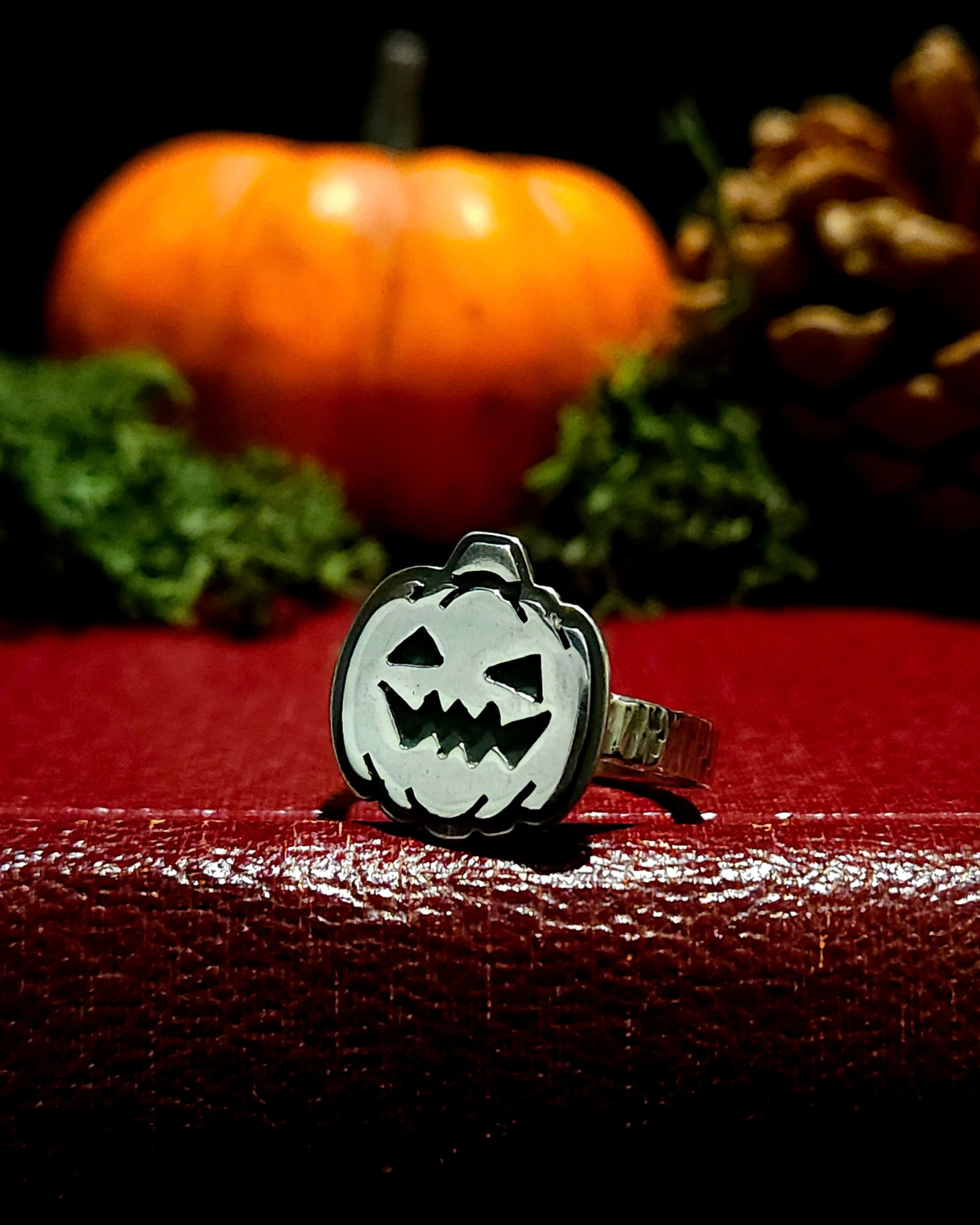 A .925 silver halloween jack-o-lantern ring sits facing the camera amongst gothic, mystical halloween staging. The face is flat, 2D with blackened details, and the band is rectangular and hammered.
It sits on a vintage red book with an orange pumpkin, pinecone and moss behind it, out of focus, all against a black background.