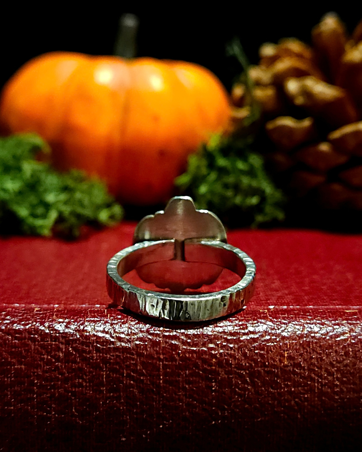 A .925 silver halloween pumpkin shaped ring sits facing away from the camera amongst gothic, mystical halloween staging. It is facing this way to demonstrate how it's made - the face is flat, 2D and the band is rectangular and hammered.
It sits on a vintage red book with an orange pumpkin, pinecone and moss behind it, out of focus, all against a black background.