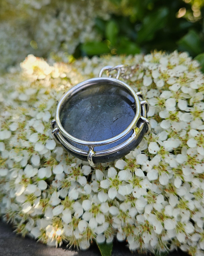 Circular labradorite & sterling silver necklace lays face down on white flowers with green leaves. The back of the pendant is smooth and flat and the image shows that the .925 pendant is that of a gallery-style claw setting.