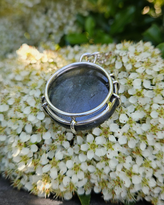 Circular labradorite & sterling silver necklace lays face down on white flowers with green leaves. The back of the pendant is smooth and flat and the image shows that the .925 pendant is that of a gallery-style claw setting.