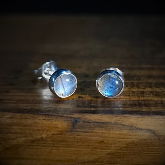A pair of round sterling silver & labradorite stud earrings on a wooden surface. Their characteristic, luminescent, blue flash is visible in both earrings.