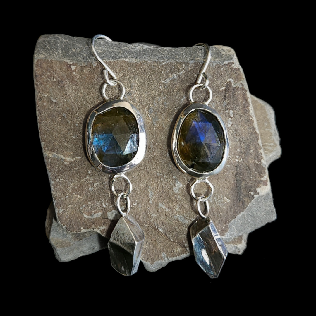 A pair of sterling silver and rose-cut faceted dangly earrings rest on a piece of slate. Their geometric and rough-cut drops underneath the gemstone are inspired by the slate mines and mountains of Wales.
