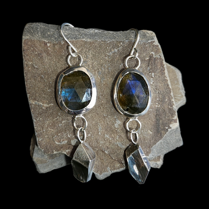 A pair of sterling silver and rose-cut faceted dangly earrings rest on a piece of slate. Their geometric and rough-cut drops underneath the gemstone are inspired by the slate mines and mountains of Wales.