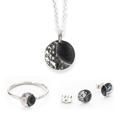 A set of lunar sterling silver jewellery including 2 rings, a pair of stud earrings, and a necklace, featuring a textured and smooth finish with a moon motif.