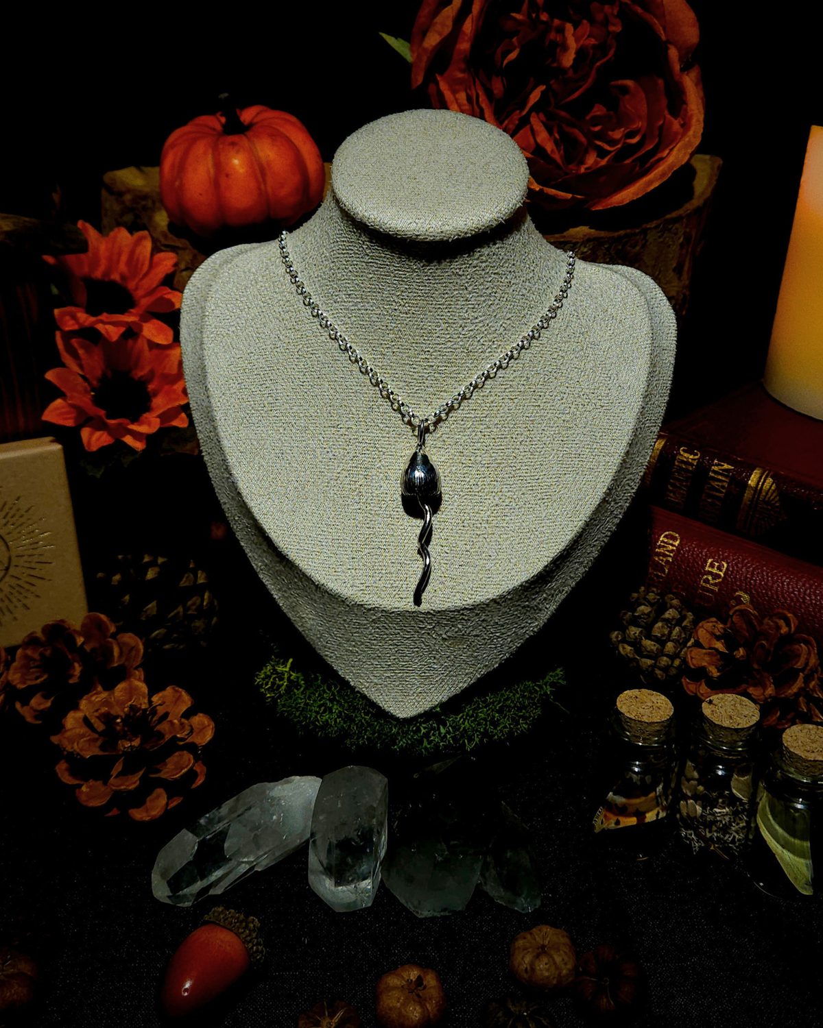 A silver chain with a solid silver magic mushroom pendant displayed on a vintage style cream jewellery bust. The photograph is styled in a witchy, moody setting with candles, pumpkins, bones, & other autumnal botanicals around it.