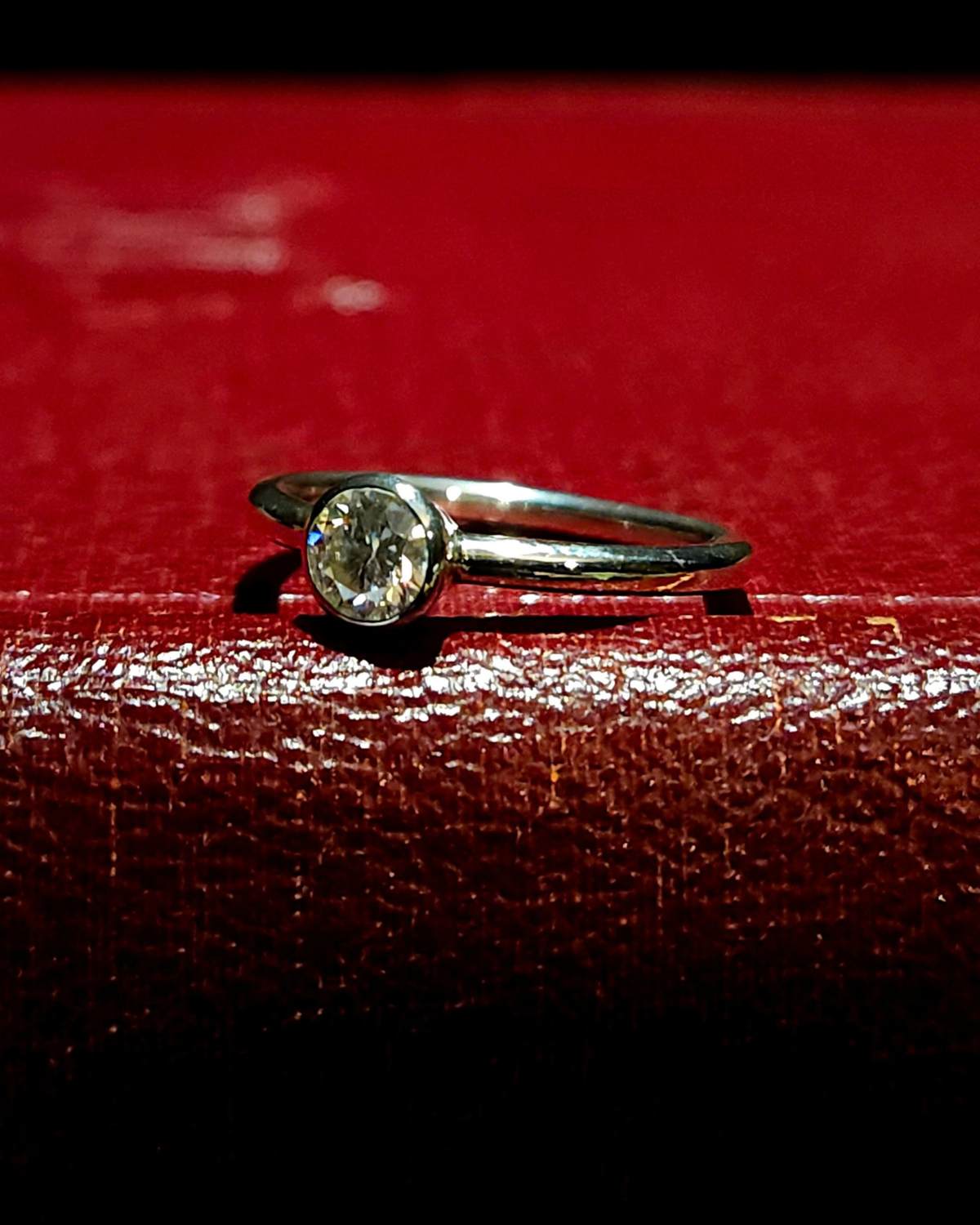 A sterling silver and moissanite solitaire ring laying on its side, stone facing slightly to the left to catch the light sparkling brightly. It lays on a dark red, vintage book.