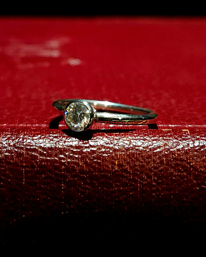 A sterling silver and moissanite solitaire ring laying on its side, stone facing slightly to the left to catch the light sparkling brightly. It lays on a dark red, vintage book.