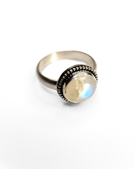 A silver ring featuring a bezel-set rainbow moonstone with a beaded border and a frosted effect on the band. The details around the moonstone have been oxidised black and then polished back to bright shining silver, leaving the recesses black to add texture and depth. The moonstone is a 10mm circle and you can see a large, bright, blue iridescent flash just to the right of centre. The background is plain white.