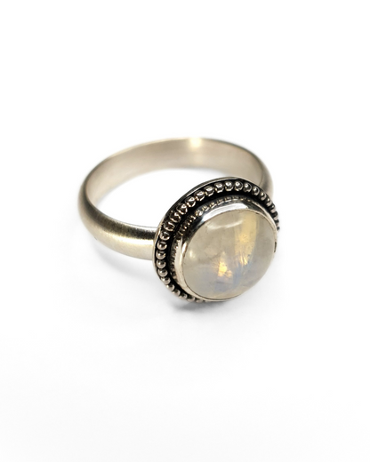 A silver ring featuring a bezel-set rainbow moonstone with a beaded border and a frosted effect on the band. The details around the moonstone have been oxidised black and then polished back to bright shining silver, leaving the recesses black to add texture and depth. The moonstone is a 10mm circle and you can see its beautiful iridescence and inclusions in the photograph. The background is plain white.