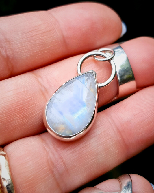 A teardrop shaped rainbow moonstone pendant is resting on an open hand to demonstrate size. The metal is bright shiny silver and the bail loop is large and sits atop it like a silver halo. The moonstone has blue irridescent flashes throughout.