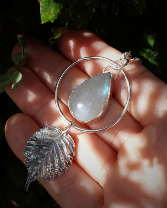 A silver pendant lays in an open palm to demonstrate its size. It is comprised of a large teardrop-shaped rainbow moonstone bezel-set in silver and surrounded by a silver halo-like circlet. A solid silver bramble leaf hangs rom the bottom of the circlet. Everything is attached by tiny jump rings so it can move naturally. The leaf is oxidised and polished to reveal the natural details including veins. The pendant is illuminated in natural sunlight showing the iridescent play of rainbows in the moonstone.