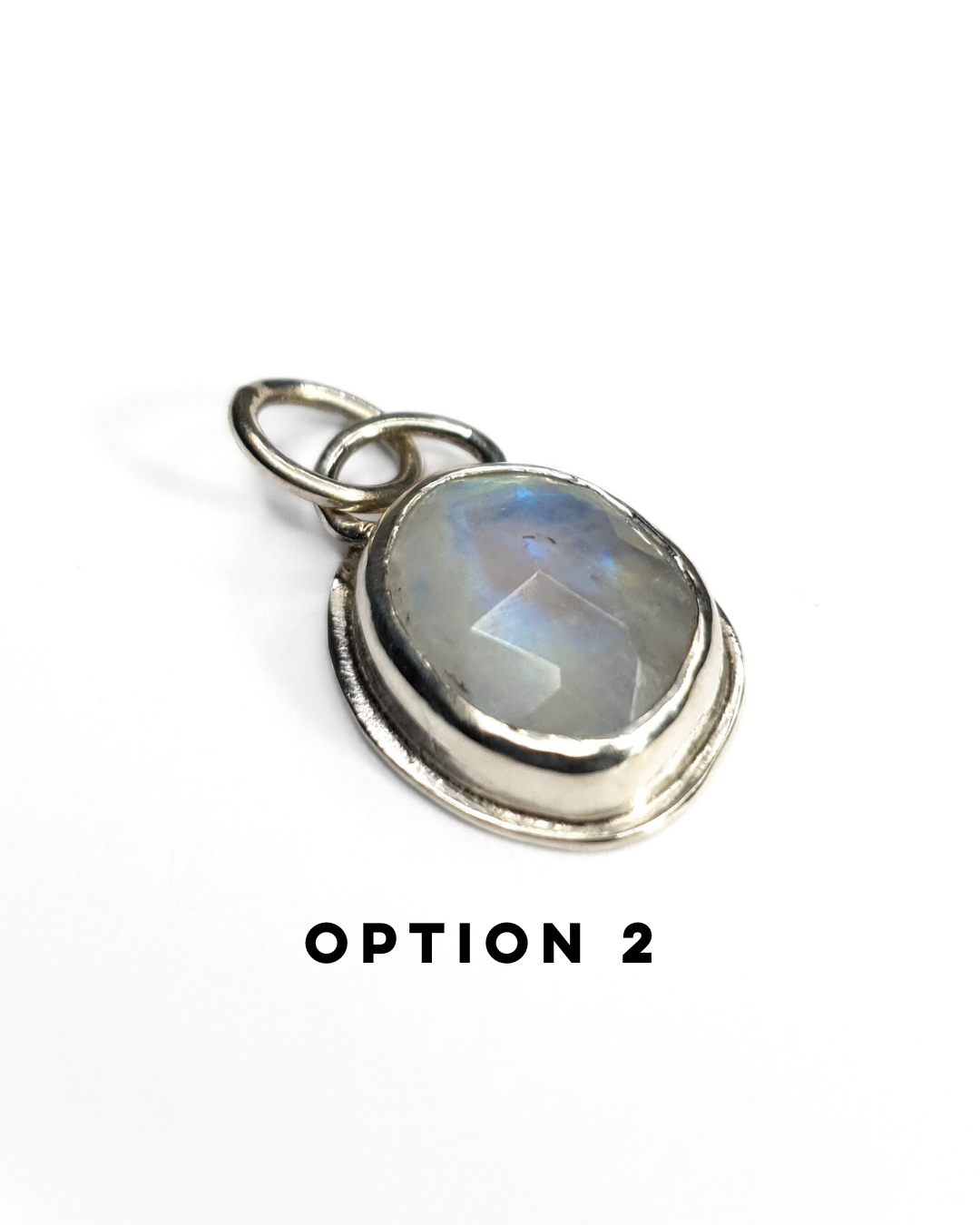 An irregular, oval-shaped rainbow moonstone is bezel set in silver and sits on a white background. The gemstone is rose-cut and the facets shine in the light displaying a beautiful show of blue-ish rainbow iridescence. There is a large circular silver bail at the top with a similarly large jump ring attached. There is text below which reads "OPTION 2"