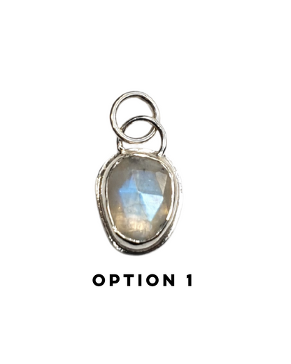 An irregular, oval-shaped rainbow moonstone is bezel set in silver and sits on a white background. The gemstone is rose-cut and the facets shine in the light displaying a beautiful show of rainbow iridescence. There is a large circular silver bail at the top with a similarly large jump ring attached. There is text below which reads "OPTION 1"