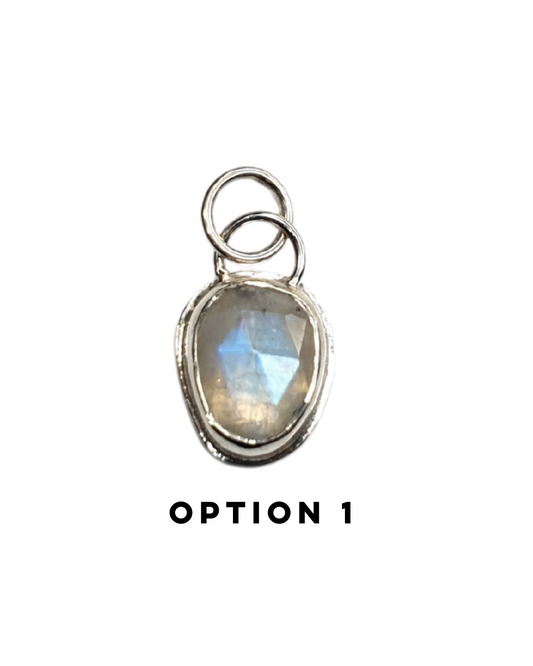 An irregular, oval-shaped rainbow moonstone is bezel set in silver and sits on a white background. The gemstone is rose-cut and the facets shine in the light displaying a beautiful show of rainbow iridescence. There is a large circular silver bail at the top with a similarly large jump ring attached. There is text below which reads "OPTION 1"