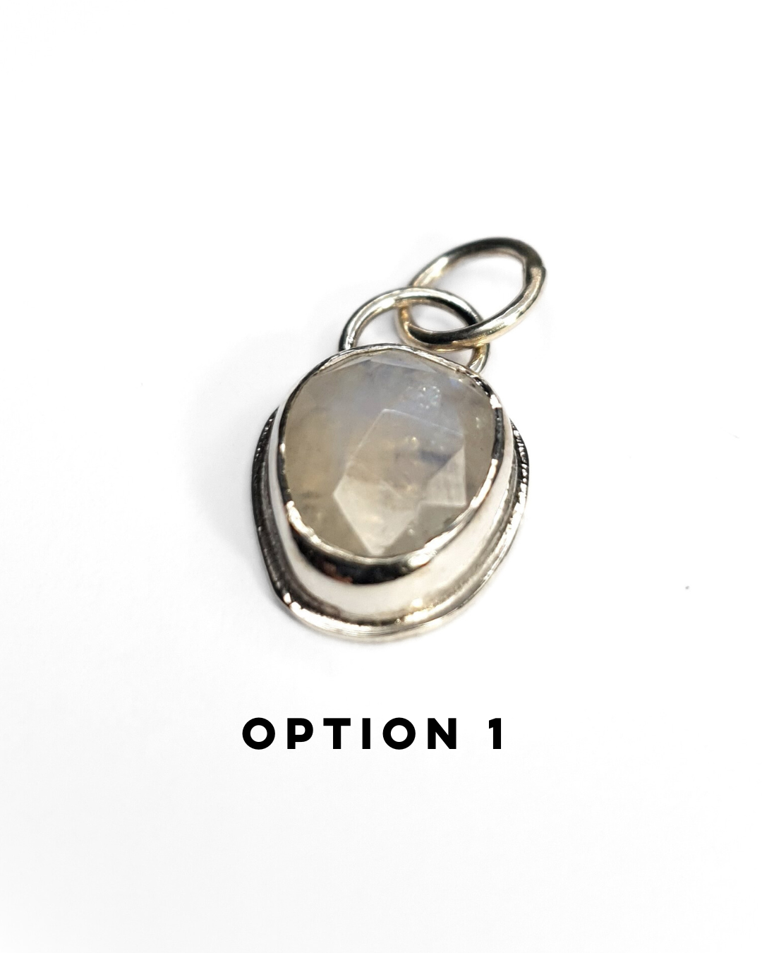 An irregular, oval-shaped rainbow moonstone is bezel set in silver and sits on a white background. The gemstone is rose-cut and the facets shine in the light displaying a beautiful show of rainbow iridescence. There is a large circular silver bail at the top with a similarly large jump ring attached. There is text below which reads "OPTION 1"