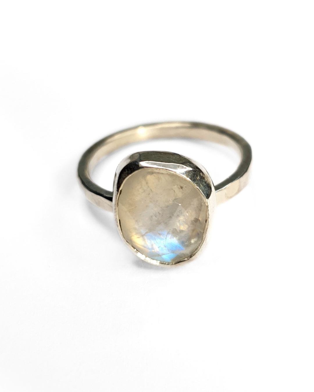 A freeform rainbow moonstone ring with a bezel setting on a shiny, hammered sterling silver band. The gemstone is faceted and contains a gorgeous iridescence and inclusions. A large blue flash is visible at the bottom. The background of the photo is white.