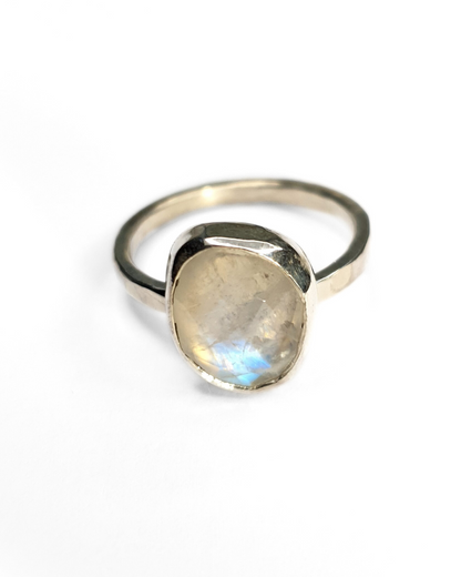 A freeform rainbow moonstone ring with a bezel setting on a shiny, hammered sterling silver band. The gemstone is faceted and contains a gorgeous iridescence and inclusions. A large blue flash is visible at the bottom. The background of the photo is white.