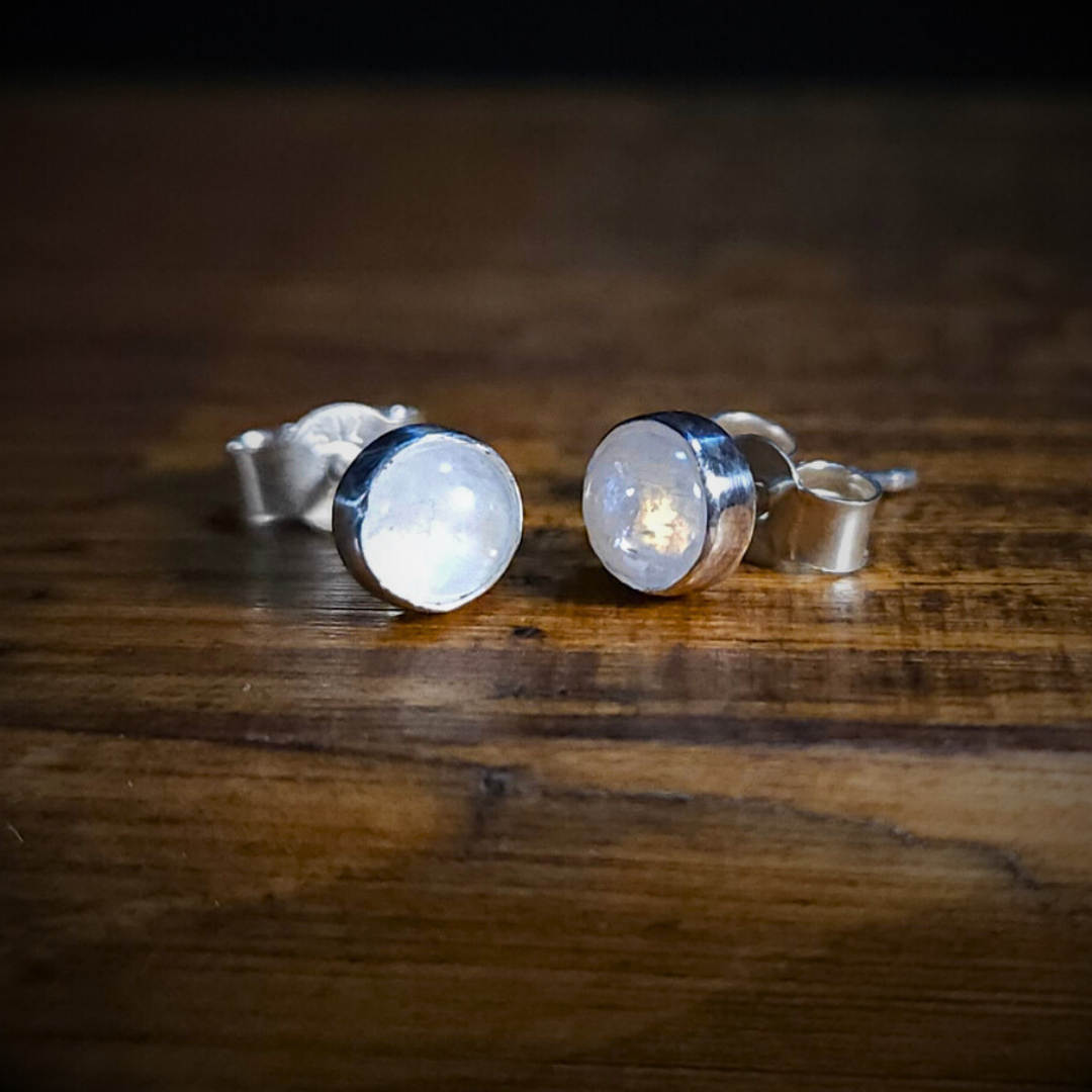 A pair of round sterling silver & rainbow moonstone stud earrings on a wooden surface. Their characteristic, luminescent play of light is visible within them - particularly clearly in the right one.