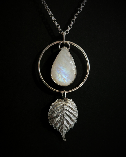 A necklace hangs on a black background. The pendant is comprised of a large, beautiful, smooth, teardrop-shaped rainbow moonstone bezel-set in silver and surrounded by a hammered silver halo-like circlet. A highly-detailed fine silver bramble leaf hangs from the bottom of the circlet. Each part is able to move independently so it can hang & flow naturally. The leaf is oxidised and polished to reveal the natural details including veins. The pendant is illuminated in natural sunlight.