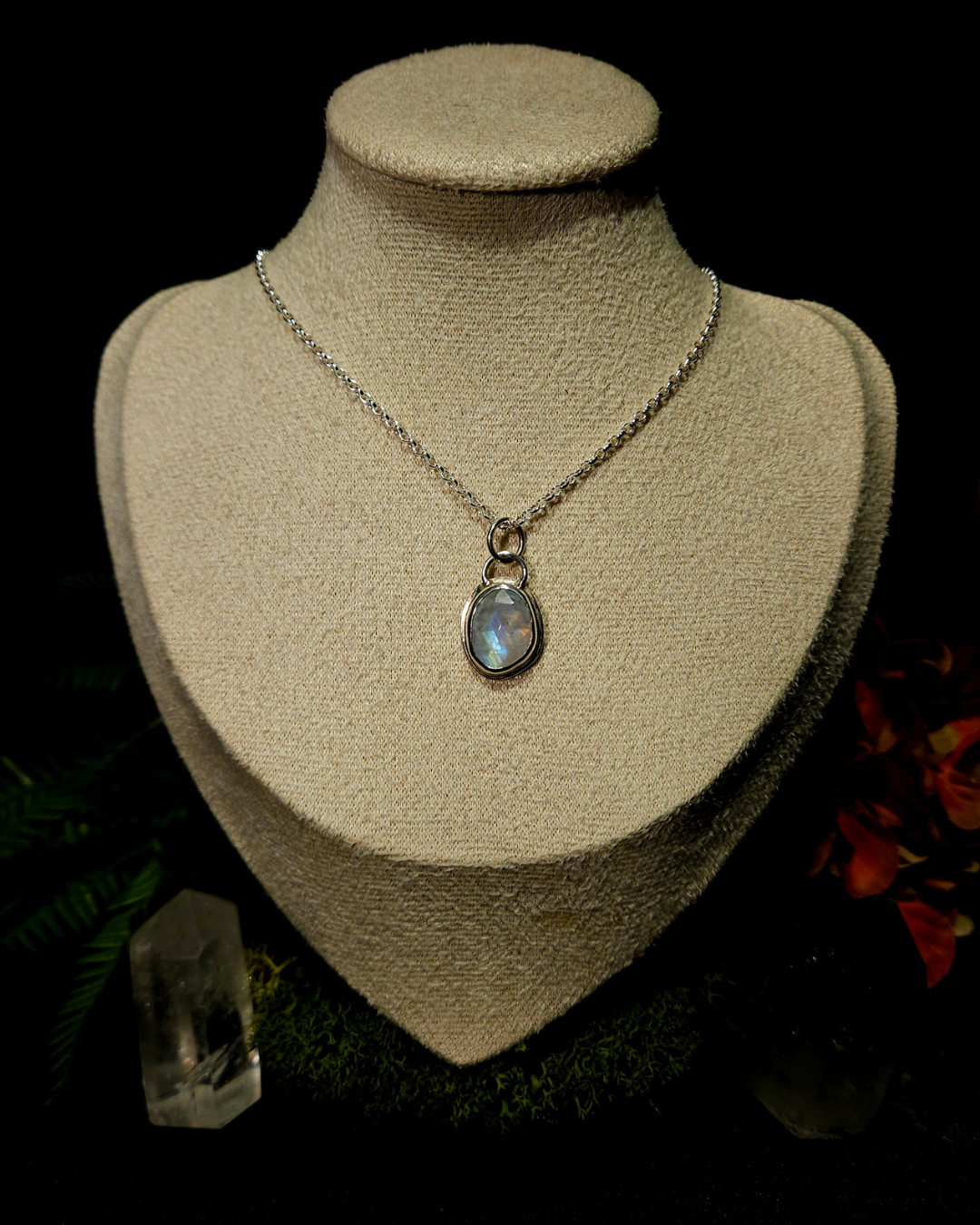 A sterling silver and glimmering rainbow moonstone necklace sit on a cream bust in a dark moody background of rain forest flowers and crystals.