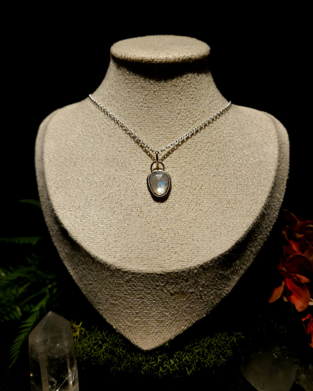 A sterling silver and lumiescent rainbow moonstone pendant on a chain displayed on a small cream bust in a dark but rich background of rainforest flowers, moss and crystals points